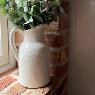 White ceramic pitcher with greenery on a brick surface