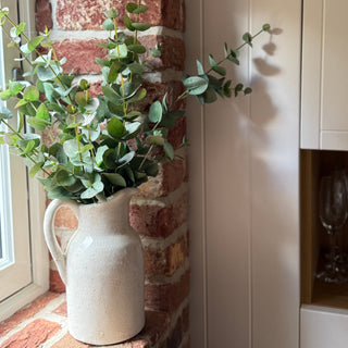 White pitcher with greenery against a brick wall and white cabinet.