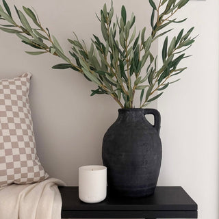 Black vase with greenery on a black table against a neutral wall.