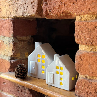 Two small white house-shaped lamps with yellow lights on a wooden shelf against a brick wall.
