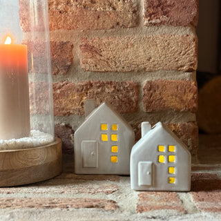 Two small ceramic house-shaped lamps with glowing interior lights on a brick surface next to a lit candle.
