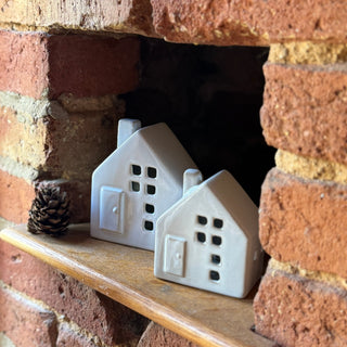 Two white ceramic house-shaped objects on a wooden shelf against a brick wall.