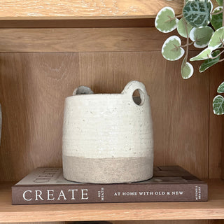 A handmade ceramic storage basket with two loop handles, featuring a textured off-white upper body and a natural light brown clay base, displayed on a book on a wooden shelf.