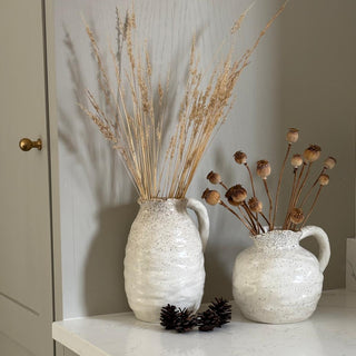 Two cream textured jug vases with dried plants on a marble surface.