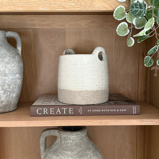 A small cylindrical ceramic planter with two loop handles sits on a book on a wooden shelf, surrounded by other ceramic vases and a green trailing plant.