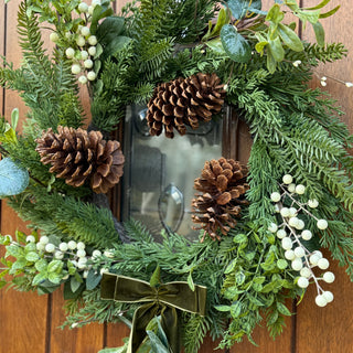 Decorative wreath with greenery, pinecones, and a bow on a wooden door.