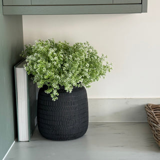 A black textured ceramic planter filled with green and white baby's breath flowers, styled on a light gray marble countertop next to a book and a woven basket, with a sage green cabinet in the background.