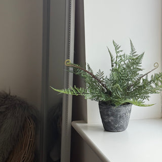 An artificial green fern plant in a dark gray textured stone pot, placed on a white windowsill with a window frame visible on the left.