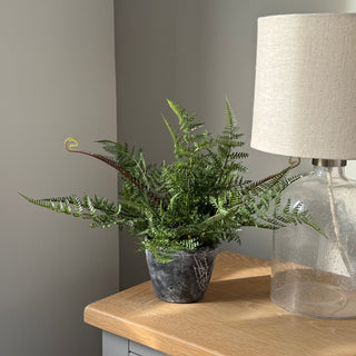 An artificial green fern plant in a dark gray textured pot, styled on a light wood side table next to a glass table lamp with a beige shade.