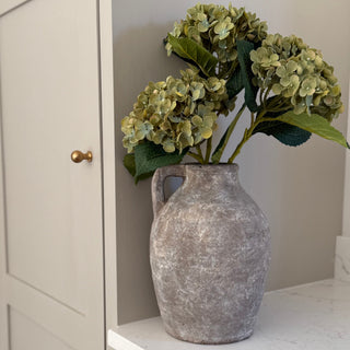 Decorative stone grey vase with green hydrangeas on a white surface next to a cabinet.
