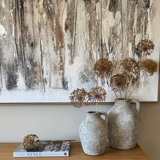 Two rustic gray ceramic jugs filled with dried brown flowers, styled on a light oak wooden console table next to a large textured wood panel wall art piece.