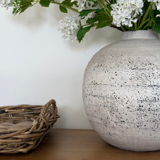 Speckled ceramic vase on a white surface with greenery and a woven object.