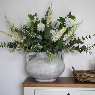 A faux flower arrangement featuring white and green colors, placed in a stone vase on a white cabinet.