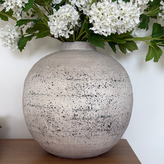 Round speckled gray vase on a wooden shelf with white flowers and green leaves.