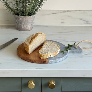 A round wooden and gray stone bread board with two slices of rustic bread on it, styled on a white marble countertop with a potted rosemary plant in the background.
