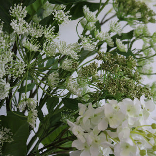 A close-up of a artificial floral display featuring white and green colors, with a variety of flowers and green leaves, arranged in a medium-sized vase.