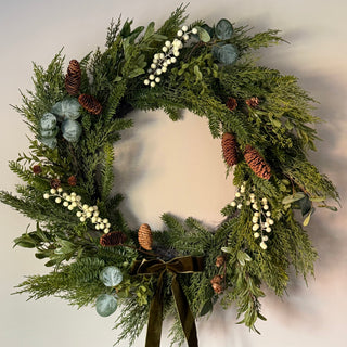 A festive green wreath decorated with pinecones, white berries, eucalyptus leaves, and a dark olive green ribbon bow at the bottom, shown against a plain light gray wall.