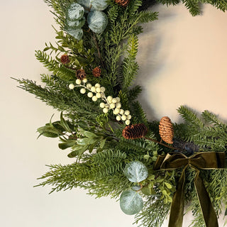 close up of a Large faux fir tree wreath with white berries, pine cones and a dark green velvet bow 