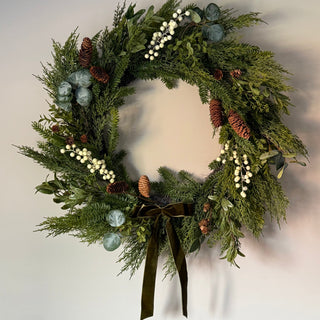 A festive green wreath decorated with pinecones, white berry clusters, eucalyptus leaves, and a dark olive green ribbon bow at the bottom, hanging against a plain light wall.