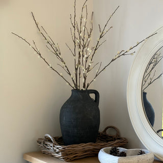 A dark textured ceramic pitcher vase filled with dried willow branches, styled on a wicker tray on a light wood console table next to a white bowl and an oval mirror.