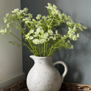 White pitcher with greenery on a woven tray against a gray wall