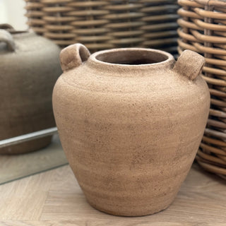 Brown ceramic pot with two handles on a wooden surface, with wicker baskets in the background.