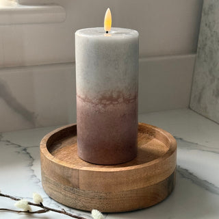 A lit pillar candle with a white-to-rose gold gradient finish sits in a round wooden holder on a marble countertop.