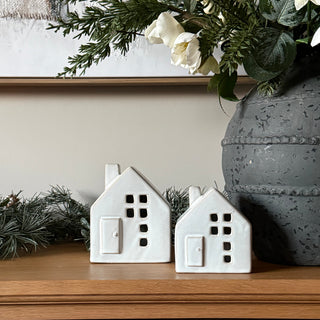 Two small white house-shaped ceramic ornaments or candle holders displayed on a wooden surface next to green garland and a gray textured vase.
