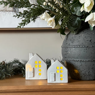 Two small white house-shaped decorative lights with yellow window accents, shown on a wooden surface next to a gray pot and white flowers.