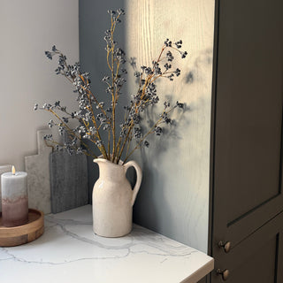 A cream-colored ceramic pitcher vase filled with  blue-gray berry branch, styled on a white marble countertop next to a lit candle and a dark wood cabinet.