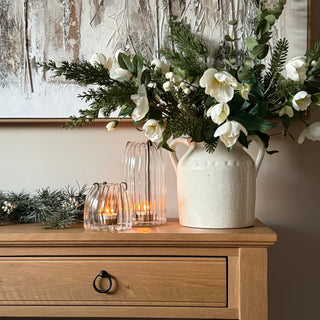 A styled holiday entryway scene featuring a large white ceramic pitcher vase filled with white flowers and green foliage, set on a light wood console table next to two lit glass candle holders and a garland.