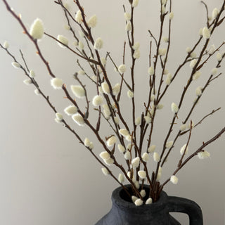 Artificial willow branch arrangement with white fluffy catkins in a dark textured ceramic vase against a plain light gray background.