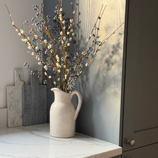 A beige ceramic pitcher vase filled with dried willow branches and small white berries, styled on a white marble countertop in a modern kitchen setting.