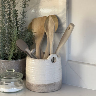 A handmade ceramic kitchen utensil holder with a white and beige glaze, filled with wooden spoons and a metal spatula, set on a kitchen counter next to a potted rosemary plant.