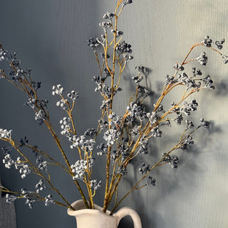 faux blue berry branch stems arranged in a white ceramic pitcher, set against a textured gray wall with a lit candle partially visible at the lower right.