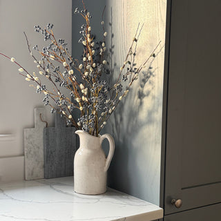 A ceramic pitcher vase filled with faux pussy willow branches and small blue berry stems, styled on a white marble countertop next to dark gray cabinets.