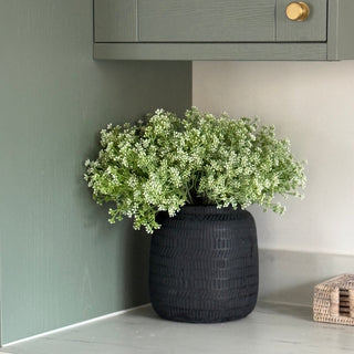 A black woven textured ceramic vase filled with green and white baby's breath flowers, styled on a light gray marble countertop in a sage green kitchen setting.