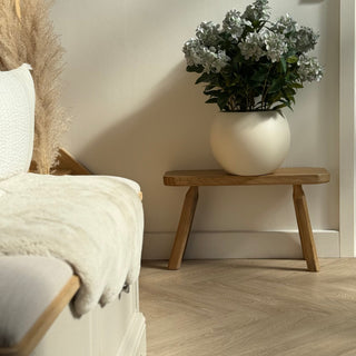 Cream vase with flowers on a wooden stool next to a white armchair in a room.