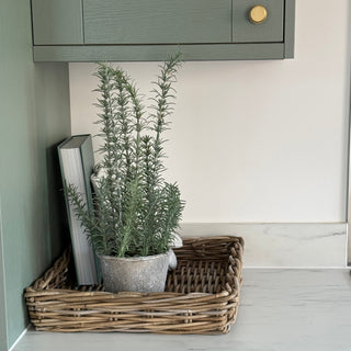 A natural woven wicker basket tray sits on a white marble countertop, holding a potted green rosemary plant and a gray book. The tray is styled in a kitchen setting with sage green cabinets and gold knobs.
