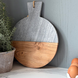 Round wooden cutting board with gray top and natural wood bottom on a marble surface with eggs and a plant.