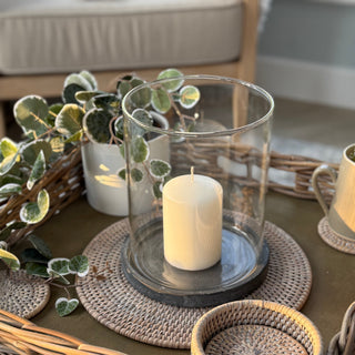 Decorative setting with a candle in a glass holder on a woven tray, surrounded by plants and a woven basket.