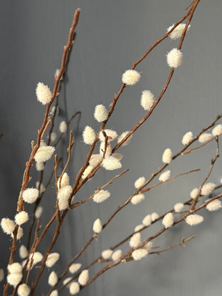 Artificial willow branches with fluffy white catkins and brown twigs, arranged against a plain gray background.