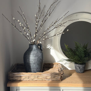 A dark gray ceramic vase filled with decorative willow branches, displayed in a woven tray on a light wood cabinet next to a round mirror and a small potted fern.