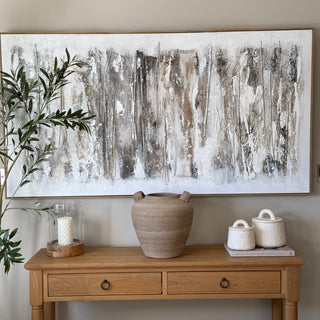 A large framed abstract canvas print with vertical brushstrokes in white, gray, taupe, and brown, displayed on a beige wall above a wooden console table with a ceramic vase and small white containers.