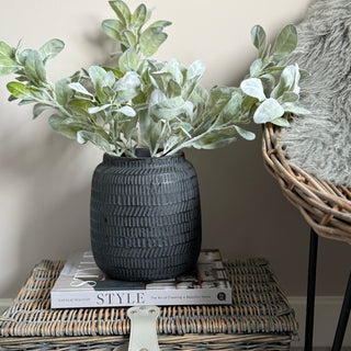Decorative scene with a textured black vase holding greenery on top of books, placed on a wicker surface.