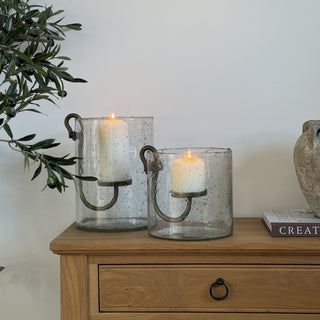 Two glass candle holders with candles on a wooden side table, next to a plant and decorative items.