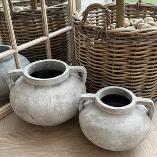 Two stone pots with handles on a wooden floor, with wicker baskets in the background.