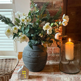 Decorative setup with flowers in a vase, candle in a glass holder, and small house-shaped light on a stone brick surface.