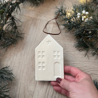 White ceramic house ornament held by a hand on a wooden surface with Christmas decorations.