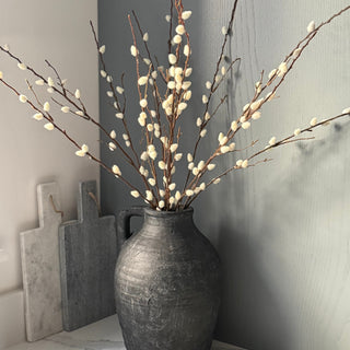 Artificial willow branches with small white buds arranged in a dark textured ceramic vase, set on a marble surface against a gray wall.